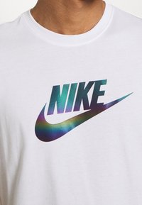 Vit t-shirt med en stor, iriserande "NIKE"-logotyp i mörkblå och gröna nyanser, kompletterad med en swoosh-design nedanför.