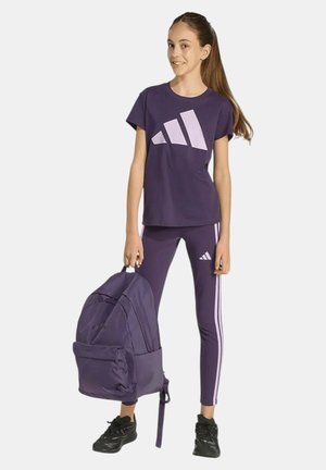 Adolescente con camiseta morada de Adidas y leggings con rayas, sosteniendo una mochila grande morada, de pie y sonriendo.