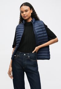 Žena s kovrčavom kosom nosi mornarsko plavi kapsi prsluk preko crne rebraste majice i tamnoplavih traperica, pozira s jednom rukom na kuku.