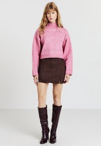 Rosa gestrickter Pullover mit hohem Kragen, kombiniert mit einem braunen Wildlederrock mit seitlichem Schlitz. Hohe, glänzende bordeauxrote Stiefel vervollständigen den Look.