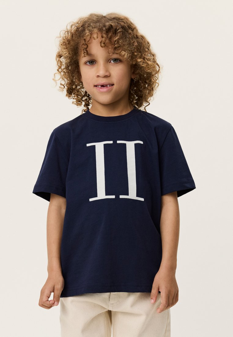 Μπλε ναυτικό βαμβακερό t-shirt με μεγάλο λευκό "II" τυπωμένο στο μπροστινό μέρος. Κοντά μανίκια και στρογγυλή λαιμόκοψη, φορεμένο με ανοιχτόχρωμο παντελόνι.