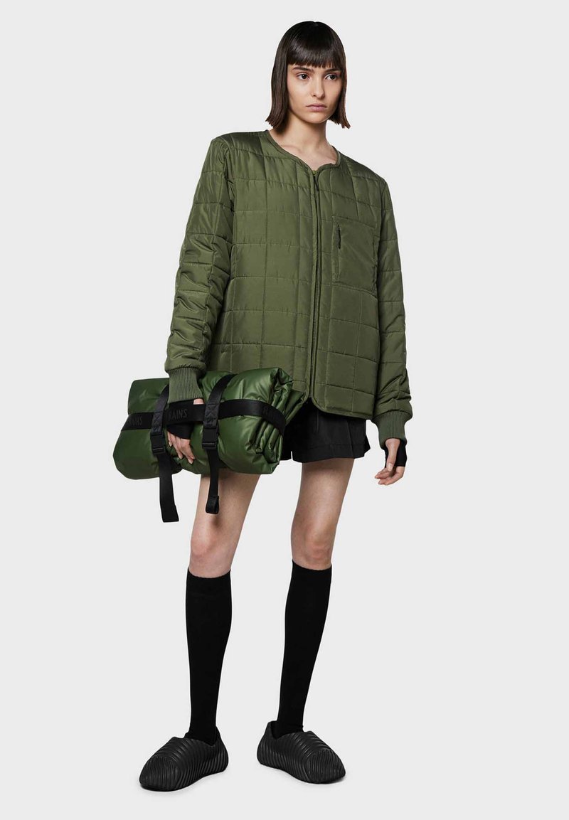 Giacca puffer verde trapuntata con zip frontale, abbinata a una borsa verde avvolta e calze nere fino al ginocchio, indossata con scarpe slip-on dalla texture.