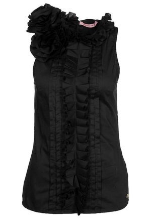 Blouse noire sans manches avec des volants verticaux sur le devant et une grande fleur en tissu sur l’épaule gauche.