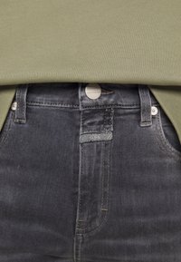 Una blusa verde oliva cae sobre unos jeans de mezclilla gris oscuro con un botón metálico y etiqueta. La tela es suave con una textura sutil y mínimos detalles de costura.