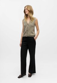 Sieviete ar gariem blondiem matiem, valkājot bezpalaku beigu krāsas žaketi ar pogām, melnas svītrainas bikses un tumšus kvadrātveida purngala apavus, stāv ar vienu roku kabatā.