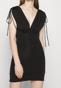 Černé přiléhavé šaty s hlubokým výstřihem do V, nařasenými rameny a nastavovacími vázáními na rukávech, vyrobené z hladké látky.