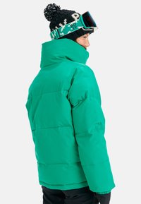 Green puffer jacket with a high collar, featuring a quilted design and a smooth texture. Worn with a black knit hat and ski goggles.