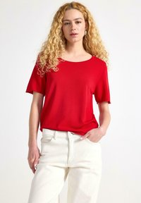 Rød kortærmet top med rund halsudskæring, lavet af glat stof, parret med højtaljede cremfargede jeans. Ingen mønstre eller pynt.