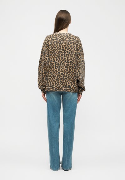 Ženska z dolgimi rjavimi lasmi, oblečena v ohlapno jopico z vzorcem leoparda in ravne modre kavbojke, stoji obrnjena stran na belem ozadju.