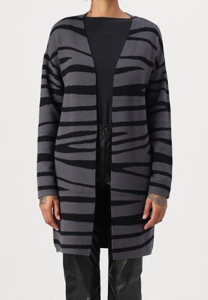 Personne portant un cardigan long gris ouvert sur le devant avec des rayures zèbre noires, un haut noir et un pantalon en cuir noir, debout les bras détendus.
