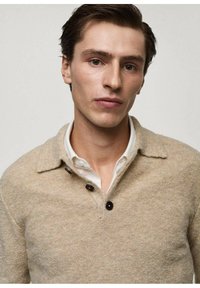 Pullover in lana beige con colletto alto, dotato di una pattina con tre bottoni e una superficie strutturata. I bottoni sono scuri e rotondi.