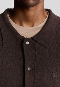Brun texturerad pikétröja med krage, med knäppning och en liten broderad logotyp på vänster sida. Lagd över en beige skjorta.