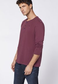 Bordeauxrotes Langarm-Henley-Shirt mit drei Knöpfen am Ausschnitt. Weicher Stoff, figurnaher Schnitt, kombiniert mit dunkelblauen Jeans.