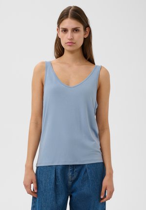 Vrouw met lang bruin haar draagt een lichtblauw mouwloos V-hals shirt en blauwe jeans, staand tegen een effen lichte achtergrond.