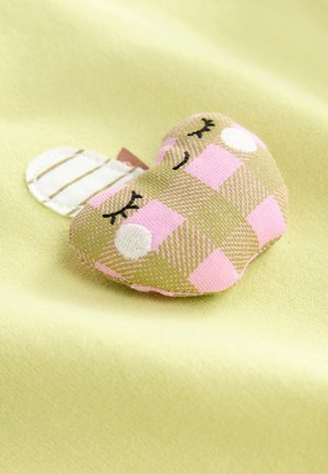 Zeleno-roza karo plišasta dekoracija v obliki srca z vezenimi obraznimi potezami, postavljena na svetlo rumeno tkanino.