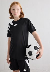 Maillot de sport noir avec des rayures diagonales blanches et un logo, associé à un short noir. Le joueur tient un ballon de soccer noir et blanc.