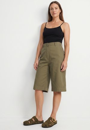 Kobieta stoi przy białej ścianie, ubrana w czarny top, oliwkowo zielone szorty do kolan oraz pasujące oliwkowe sandały z klamrą.