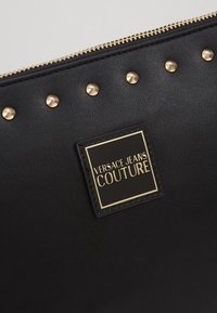 Clutch in pelle nera con borchie dorate lungo il bordo superiore, con una prominente placca quadrata con il logo che recita "Versace Jeans Couture."