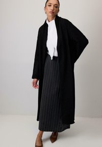 Cardigan lungo nero in tessuto testurizzato, indossato sopra una blusa bianca e una gonna a righe nere. Scarpe marroni a punta completano l'outfit.