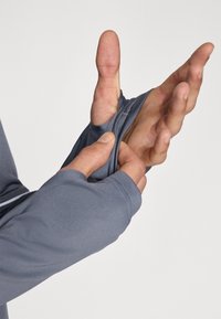 Grå langermet skjorte med tommelhull; glatt tekstur, tettsittende design og sømmer. Stoffet fremstår lett og elastisk.
