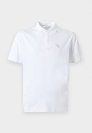 Homme debout portant un polo blanc à manches courtes, un pantalon large beige et des chaussures marron, une main dans la poche.