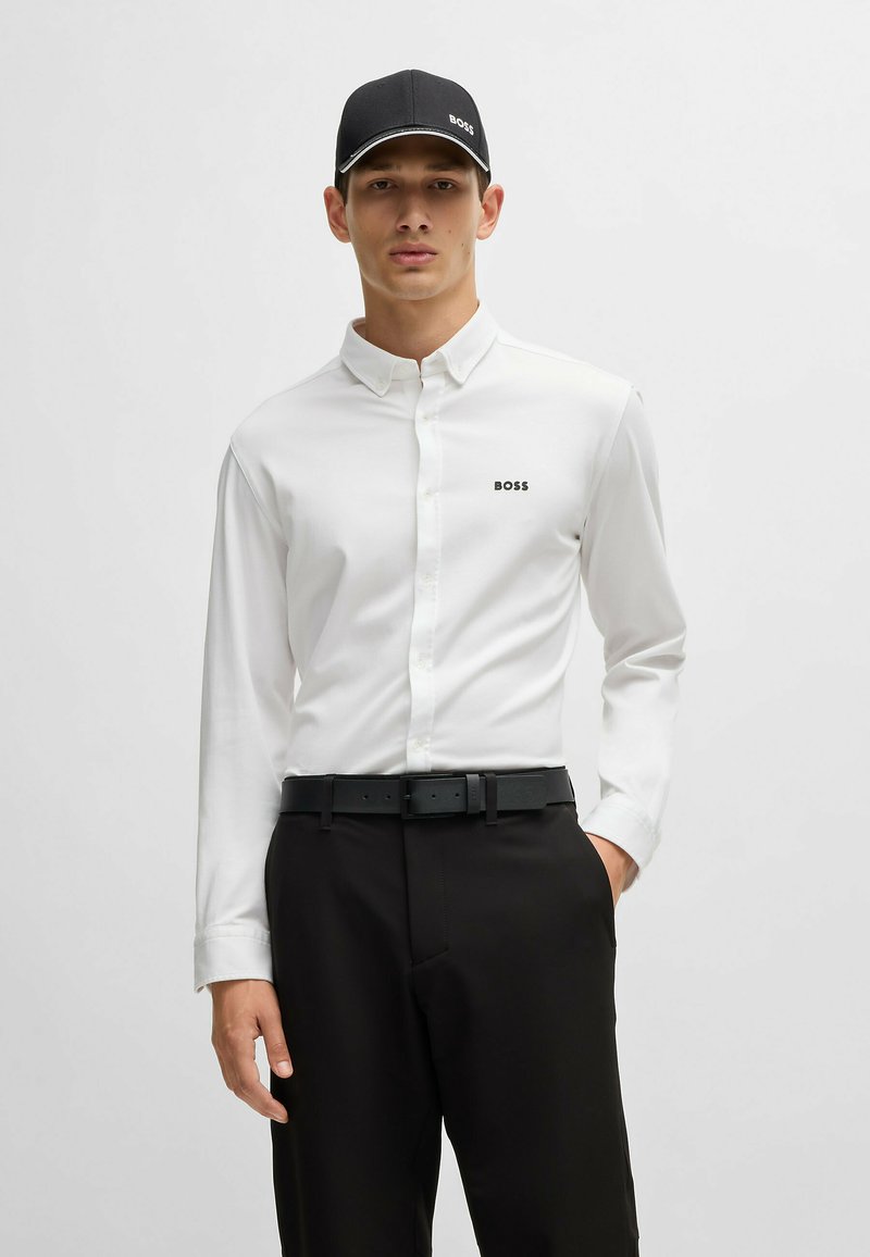 Witte button-up shirt met een klein zwart "BOSS" logo, gecombineerd met zwarte broek en een zwarte pet met een wit logo en rand.
