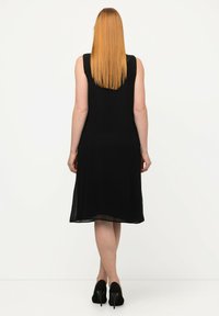 Vestido negro sin mangas con un diseño fluido, hasta la rodilla, que presenta una textura suave y una ligera transparencia en el dobladillo. Tacones negros altos.