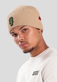 Mężczyzna w beżowej dzianinowej czapce z zieloną naszywką w kształcie lalki oraz białym swetrze z napisem "WRONGS FRIENDS" na jednolitym szarym tle.