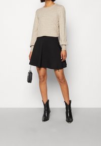 Pull en maille beige avec manches retroussées, associé à une jupe évasée noire. Bottines noires à bouts pointus. Petit sac à main noir avec chaîne.