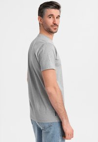 Camiseta gris de manga corta con un patrón moteado. Está hecha de una tela suave con un cuello redondo clásico y un dobladillo ligeramente más largo por detrás.