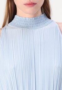 Světle modrý skládací top s vysokým nařaseným límcem a bezrukávovým designem. Hladká textilie s vertikálními pruhy a jemnými detaily na výstřihu.