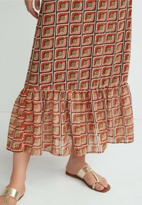 Jupe maxi avec un motif géométrique dans des tons de marron, rouge et crème. Ourlet évasé avec des volants, fabriquée en matériau léger.