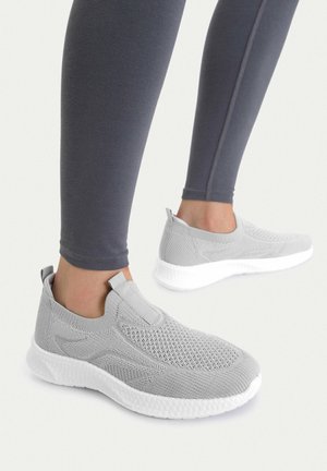 Pies con zapatillas deportivas tipo slip-on de tejido gris con suelas blancas texturizadas y leggings gris oscuro sobre un fondo liso.