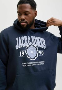 Sudadera azul marino con un gran gráfico blanco que presenta el texto "JACK&JONES 1990" y un logo circular. Material suave, bolsillo frontal, capucha ajustable.