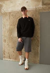 Felpa nera con colletto arancione, pantaloni corti grigi con logo, calze bianche e sneakers bianche con accenti marroni. Sfondo neutro testurizzato.