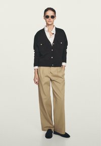 Cardigan nero lavorato a maglia con due tasche sul petto e bottoni dorati, indossato sopra una camicia bianca, abbinato a pantaloni beigi a pieghe e scarpe nere.