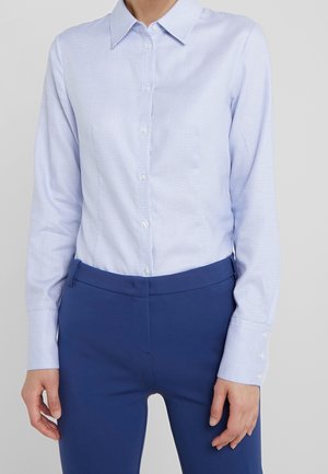 Person wearing a light blue patterned button-up shirt tucked into fitted dark blue pants, standing with hands relaxed at sides.