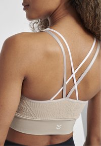 Beige sport-bh med texturerad rygg, tunna justerbara axelremmar i ljus beige och vitt, samt tydlig branding på den nedre banden.