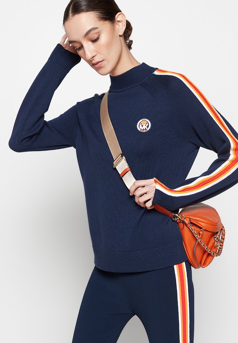 Donkerblauwe gebreide trui met oranje en gele strepen langs de mouwen, voorzien van een ronde logo patch. Gepaarde met een strakke donkerblauwe broek en een oranje tas.