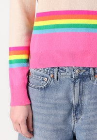 Lysebeige strikkegenser med horisontale striper i rosa, blått, grønt, oransje og gult; kombinert med lyseblå denimjeans.