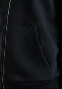 Felpa nera con zip, particolare della tasca e parte della zip, che mostrano la morbida texture del tessuto e i dettagli delle cuciture.