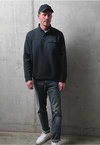 Muž v černé čepici, černém svetru s kapsou na prsou, modrých džínách a bílých teniskách stojící proti hladké betonové zdi.