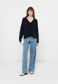Donna che indossa un maglione nero a scollatura a V, jeans larghi blu chiaro, scarpe bianche e tiene in mano una piccola borsa bianca, in piedi contro uno sfondo bianco.