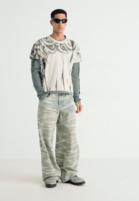 T-shirt graphique avec imprimé abstrait, superposé à un haut à manches longues. Pantalon à motifs vert clair, ample. Les accessoires comprennent des lunettes de soleil sombres.
