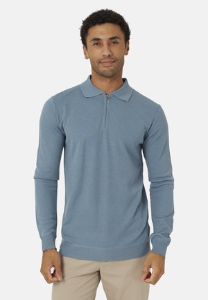 Uomo che indossa una polo a manica lunga azzurro chiaro con collo con cerniera e pantaloni beige, in piedi su uno sfondo uniforme.