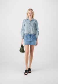 Camisa de ganga azul clara com fecho de botão e bolso, combinada com uma saia de ganga com a bainha desfiada, sandálias pretas de slide e uma mala verde.