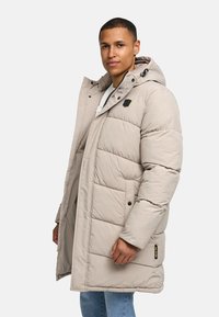 Beige puffer jacket with a hood, featuring a quilted design, black accents, a front zipper, and snap buttons. Worn over a white shirt.