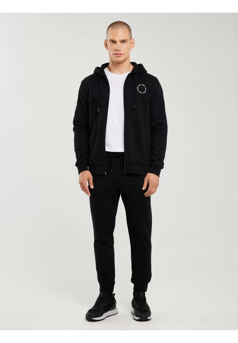 Bărbat stând, purtând hanorac negru cu fermoar și logo circular alb, tricou alb, pantaloni sport negri și adidași negri pe un fundal simplu.