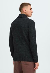 Maglione nero a maglia con colletto alto, polsini e orlo a costine, caratterizzato da un motivo testurizzato. Indossato con pantaloni marroni.