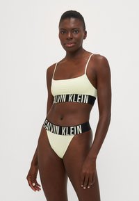 Lys gul bralette og matchende bikiniset med en svart elastisk midje med "CALVIN KLEIN"-logo. Glatt stoff, minimalistisk design.